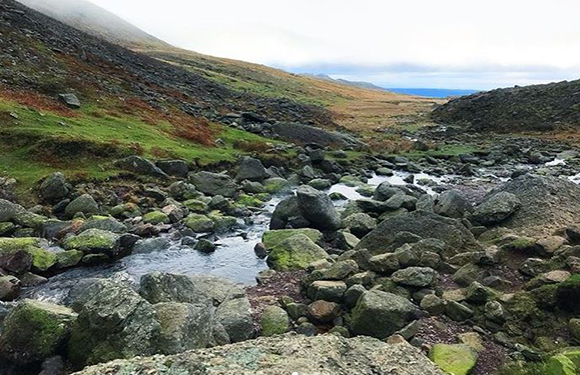 A stream flowing through a rocky landscape with grassy hillsides and a cloudy sky in the background.