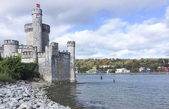 A stone castle with towers sits by a body of water, with a rocky shore upfront and trees and buildings across the water.