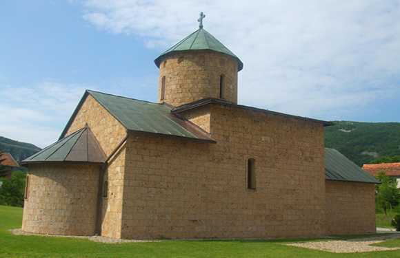 A small stone church with a green metal roof and a single bell tower, set against a backdrop of green hills and a clear sky.