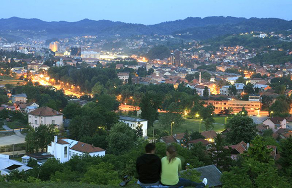 A couple sitting on a hill overlooking a cityscape at dusk with lights beginning to illuminate the buildings and streets.