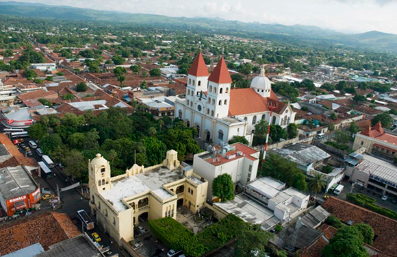 Aerial view of a town with a prominent church featuring two white spires, surrounded by trees and buildings under a cloudy sky.