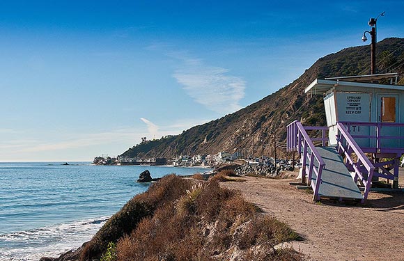 A coastal landscape with a lifeguard tower on the right, a clear blue sky above, and a mountain range extending into the distance on the left.