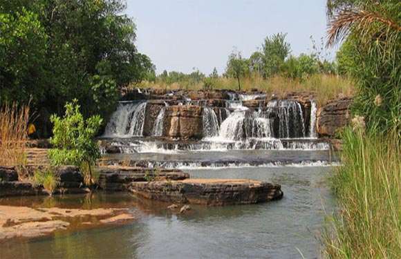 A cascading waterfall with multiple levels over rock formations, surrounded by green vegetation under a clear sky.