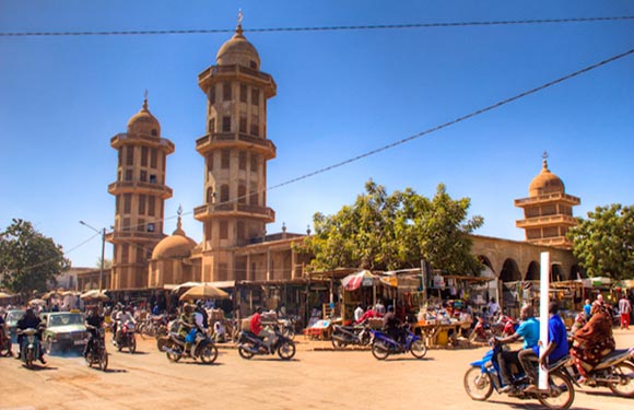 A busy street with people and motorcycles before a unique building with twin towers and a dome under a clear sky.