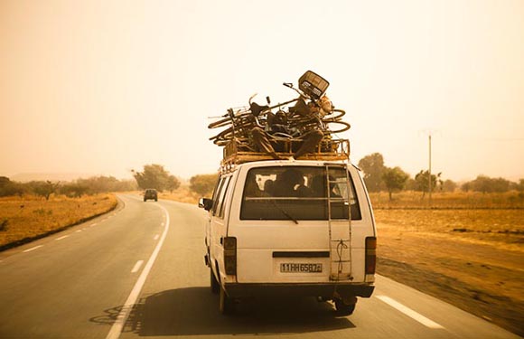 A van heavily loaded with bicycles on its roof driving on a road with a dusty, hazy sky in the background.