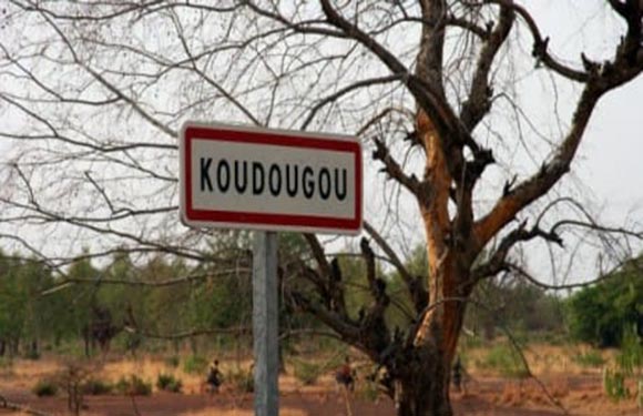 A road sign with the text "KOUDOUGOU" in front of a leafless tree and a dry landscape.