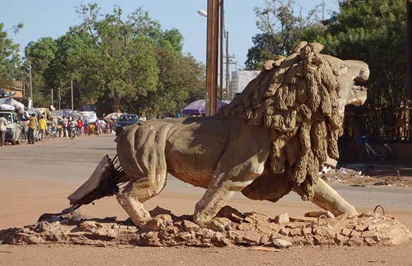 A bronze-like statue of a striding lion stands on a pedestal in a dusty street, with people and buildings behind.