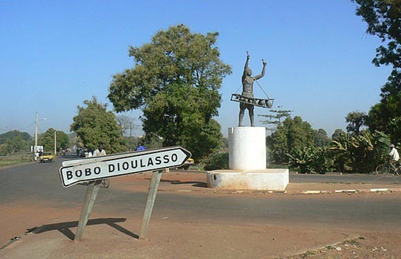 A statue with raised arms is on a roadside pedestal, with a sign pointing to "BOBO DIOULASSO" and a backdrop of trees and clear sky.