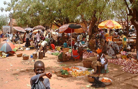 An outdoor market bustling with stalls selling fruits and vegetables, a child in the foreground, trees and clear sky in the background.