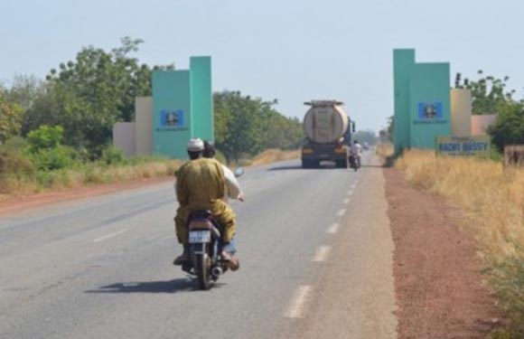 A person riding a motorcycle on a road with a large truck ahead and entrance gates to a facility on both sides of the road.