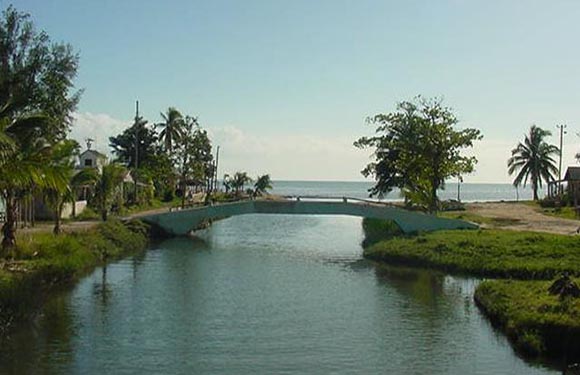 A small bridge over a narrow waterway leading to a larger body of water, with greenery on both sides and a clear sky above.