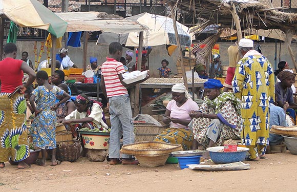 A busy outdoor market in an African setting, with people trading goods at stalls under colorful umbrellas.