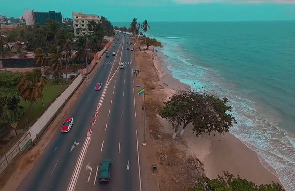 Aerial view of a coastal road with vehicles, adjacent to a beach with waves hitting the shoreline, and buildings in the background.