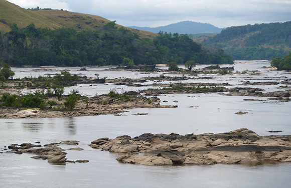 A river with multiple exposed rocks and small islets, surrounded by hilly terrain with sparse vegetation under a cloudy sky.