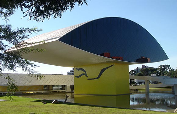 A unique modern building with a blue disc-like roof, supported by a yellow pillar, located near water and trees.