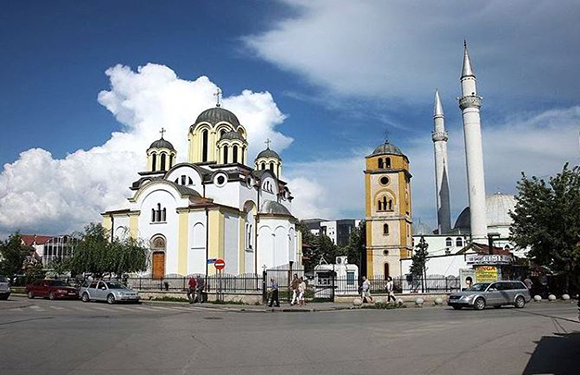 A cityscape with a golden-domed church and a tall minaret mosque, under a partly cloudy sky, bustling with people and parked cars.