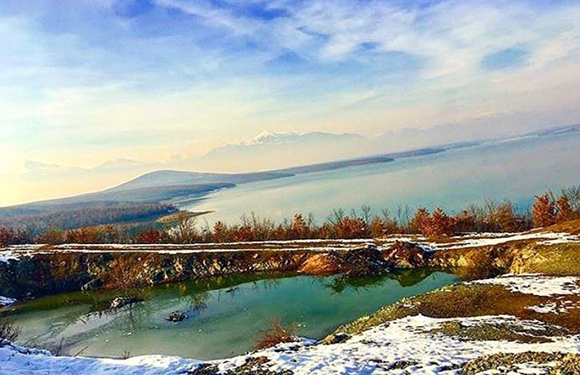 A scenic landscape featuring a tranquil lake with surrounding foliage and a mountain in the distance under a partly cloudy sky.