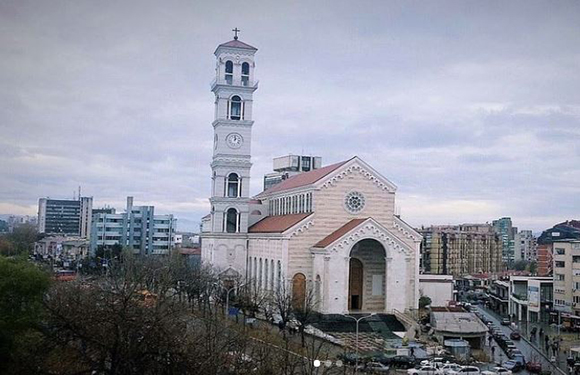 An image of a white church with a prominent bell tower, surrounded by urban buildings under an overcast sky.