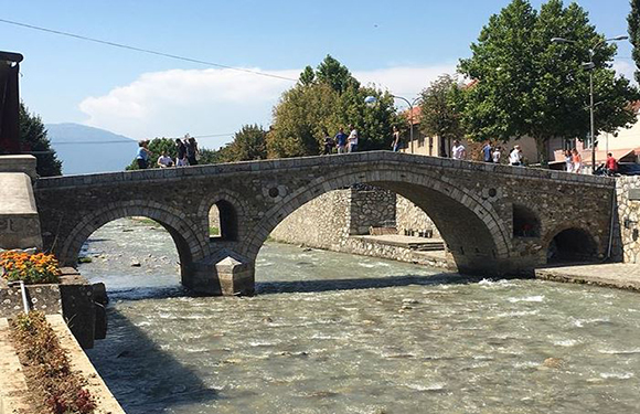 An old stone bridge with two arches over a flowing river, with people walking on and around the bridge, and trees and buildings in the background.