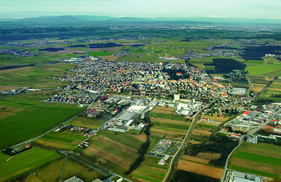 Aerial view of a small town with dense housing surrounded by green fields and sparse industrial areas under a clear sky.