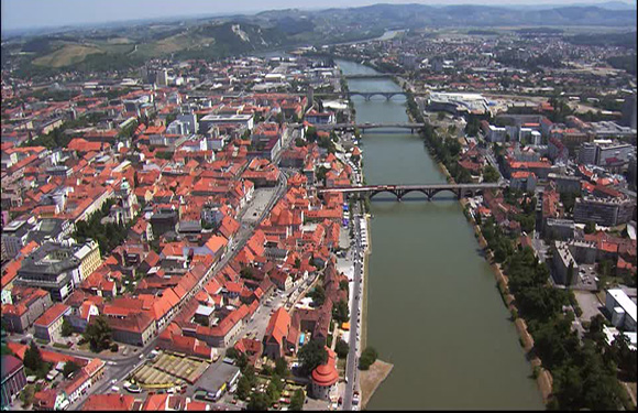 Aerial view of a city with red-roofed buildings, several bridges over a river, and greenery surrounding the urban area.