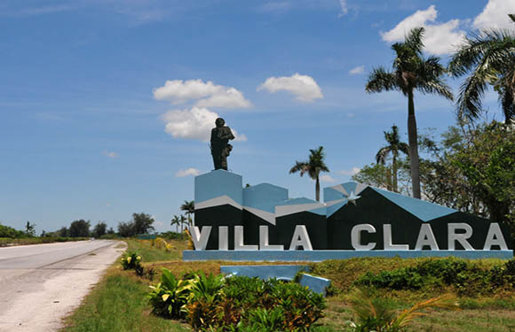 A statue on a pedestal overlooking the text "VILLA CLARA" in large letters, with palm trees and a blue sky with clouds in the background.