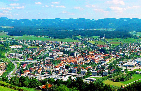 A panoramic view of a small town with dense buildings surrounded by greenery and hills under a partly cloudy sky.