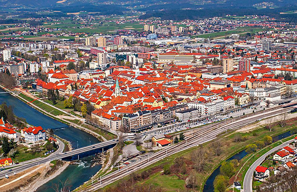 Aerial view of a city with red-roofed buildings, a river, greenery, distant hills, and a clear sky.