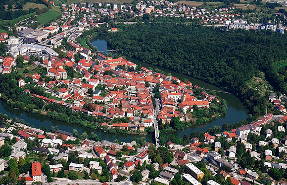 Aerial shot of a historic town with red-roofed buildings, encircled by a river, with modern structures and greenery on the outskirts.