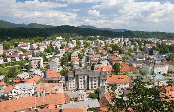 Aerial view of a small town with dense housing and buildings, surrounded by greenery and hills in the background under a partly cloudy sky.