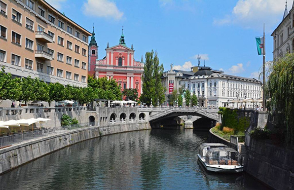 A scenic city view with a river, bridge, riverside buildings, and a pink-white church under a blue, cloudy sky.