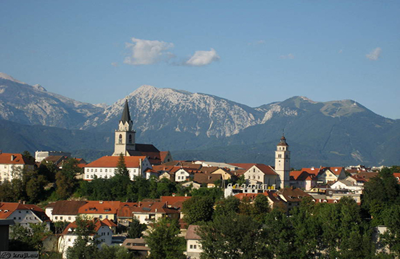 A panoramic view of a European town with traditional buildings and a church spire, set against a backdrop of distant mountains under a clear sky.