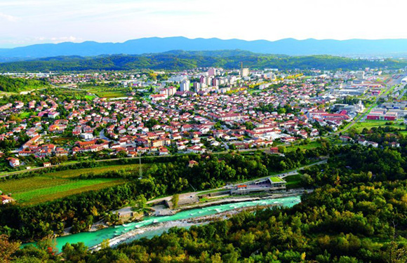 Aerial view of a town with buildings, streets, greenery, a river on the left, hills in the background, and a cloudy sky.