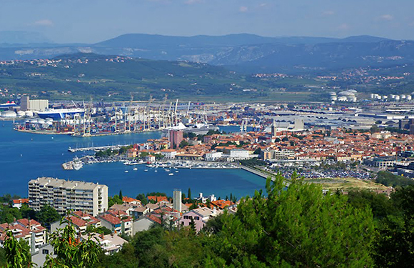 Aerial view of a coastal city with a marina, densely packed buildings, and surrounding greenery under a clear sky.