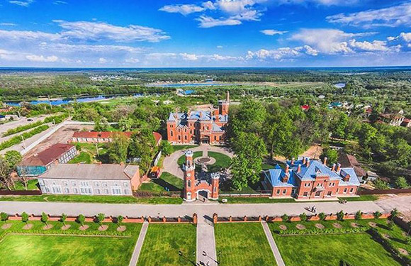 Aerial view of a grand red brick building with chimneys and towers, set amid green lawns and trees under a partly cloudy sky.