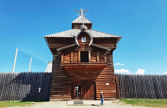 A person stands before a traditional wooden building with a dome, enclosed by a tall fence under a clear sky.