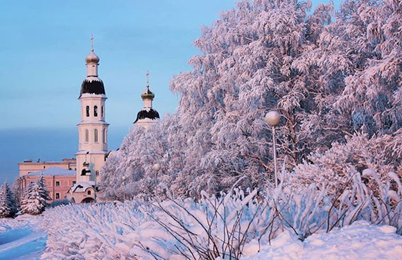 A snowy winter scene featuring snow-laden trees, a white church with golden domes, under a pastel sky.