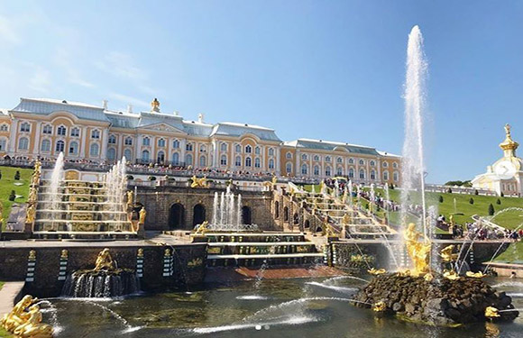 A sunny day photo of Russia's Peterhof Palace, showcasing its grand fountains, golden statues, and strolling visitors.