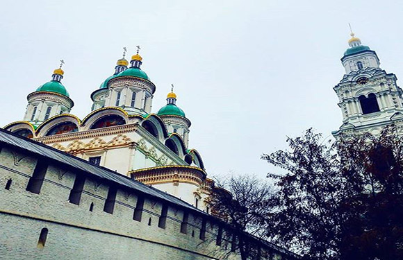 An image of a historic building with green domes and a bell tower, partially obscured by trees, behind a stone wall.