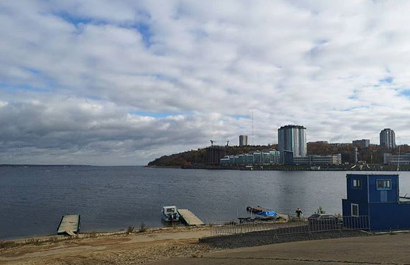 A landscape view of a waterfront with buildings in the distance, clouds in the sky, and a small boat and structures on the shore in the foreground.