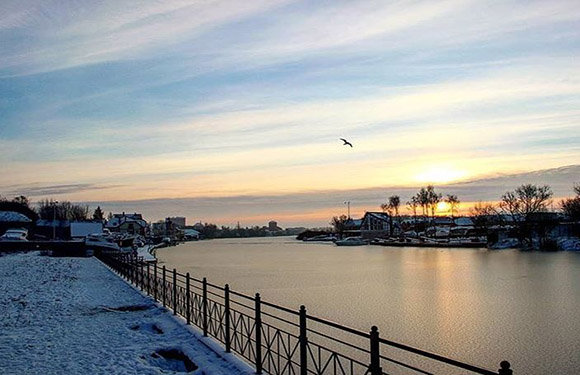 A winter sunrise over a calm river with snow-covered banks, a metal fence in the foreground, houses on the left, and a single bird in flight.