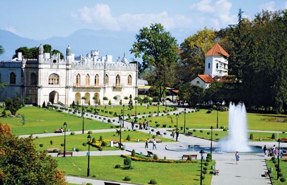 A grand white building with arches sits in a manicured park with a fountain, trees, and people relaxing, against a mountainous backdrop.