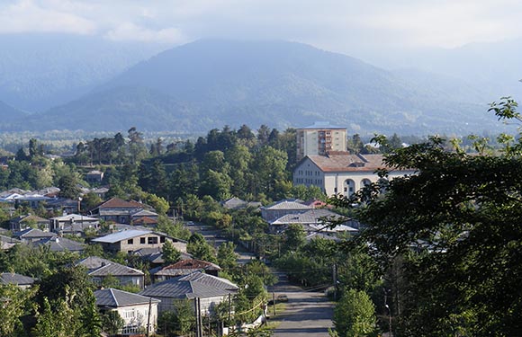 A scenic view of a hilly region with greenery, buildings in the foreground, mountains in the back, under a semi-cloudy sky.