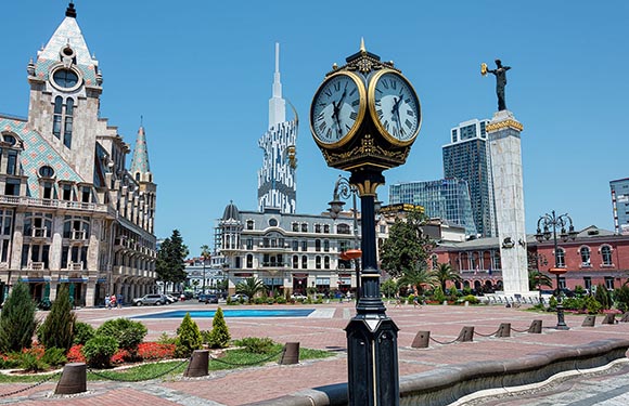 A sunny day showcases a street clock, a cityscape with a historic tower, modern skyscrapers, and a columned statue.