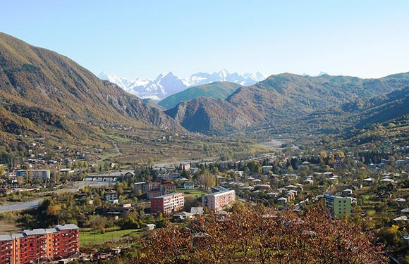 A panoramic view of a valley with a small town nestled among rolling hills and mountains in the background under a clear sky.