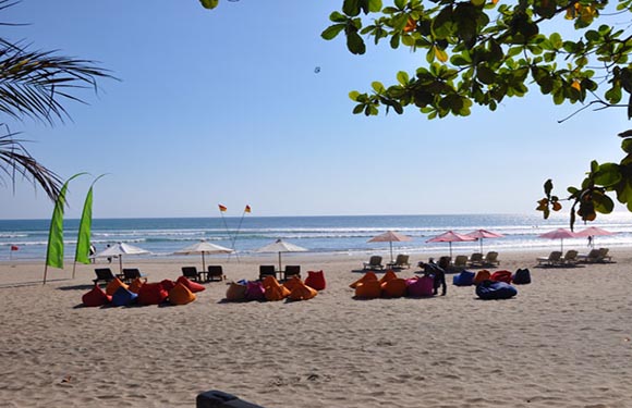 A serene beach scene with blue skies, calm sea, colorful bean bags, umbrellas, and green foliage.