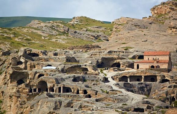 Ancient cave dwellings carved into a hillside, with a brick structure among them, set against green hills and a cloudy sky.