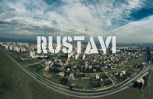 Aerial view of a cityscape with large block letters spelling "RUSTAVI" superimposed over the image.