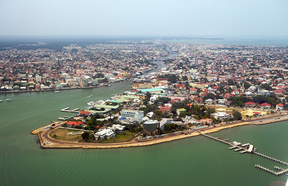 Aerial view of a coastal city with dense building clusters, roads, and green areas, surrounded by water with a clear sky above.