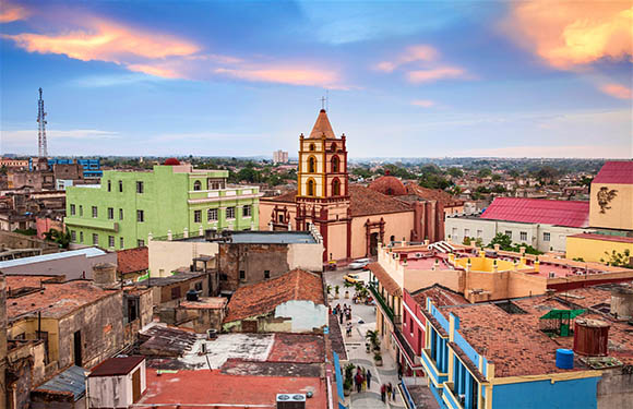 Aerial view of a colorful town at sunset with historic buildings and a prominent church tower under a sky with pink and blue hues.